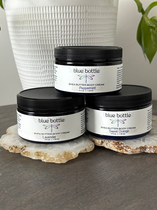 Three jars of Blue Bottle Body Care shea butter body cream on a stone surface with a white textured vase in the background.